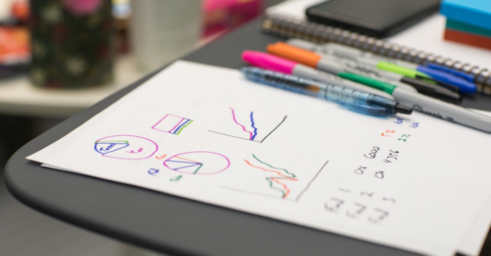 White sheet of paper with hand drawn coloured graphs lying on a desk with pens