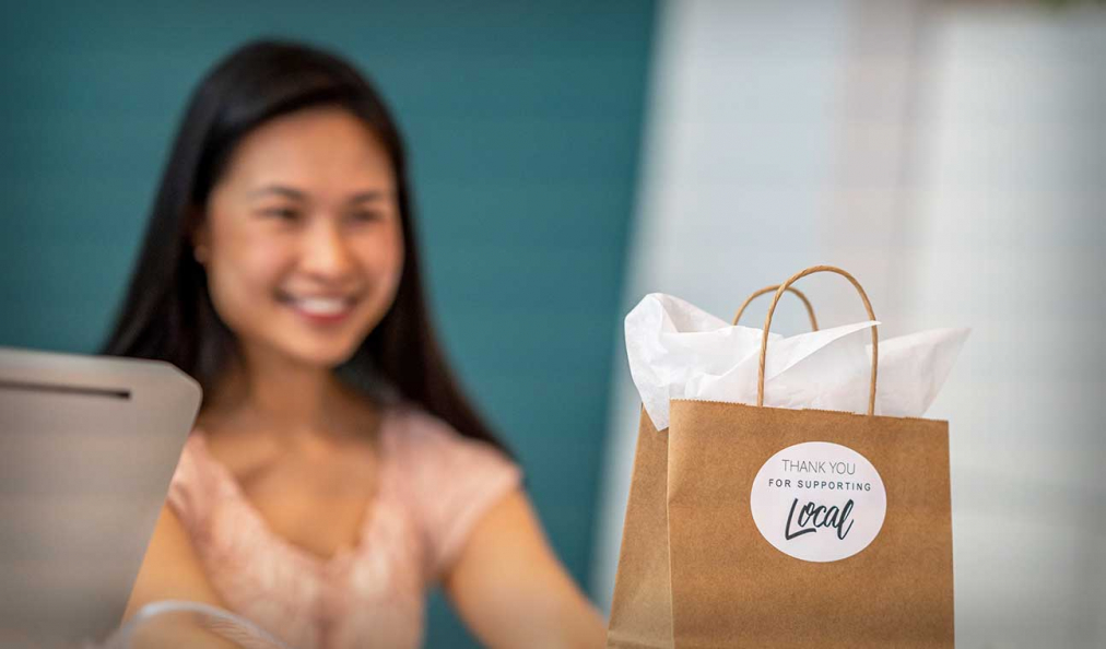 Smiling woman from a local community store offering a paper bag with thank you for supporting local