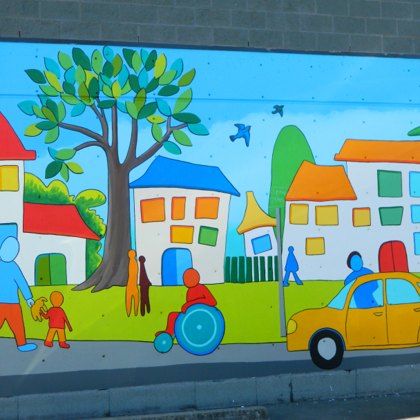 A wall mural of a residential street with a row of houses, a sidewalk with pedestrians and a person in a wheelchair and a parked car in front