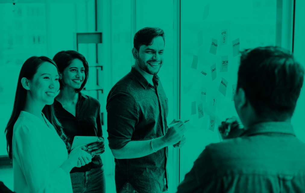 Four colleagues smiling and discussing ideas around a glass wall covered in sticky notes during a collaborative planning session.