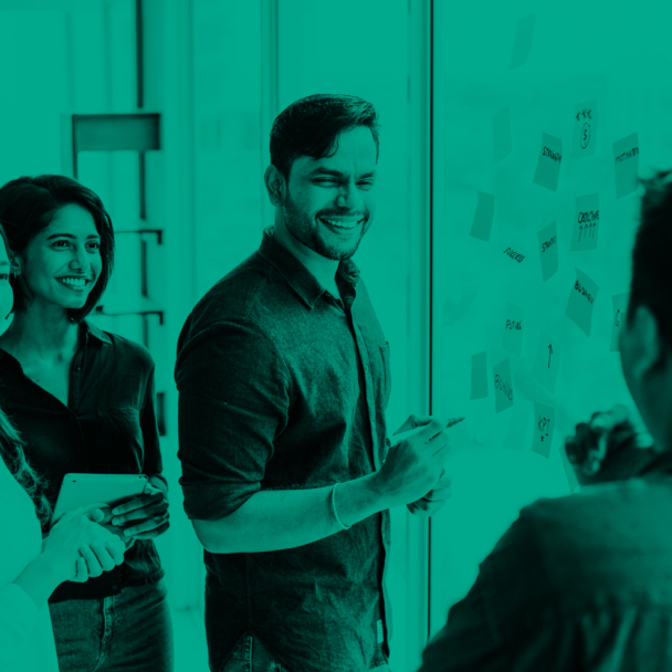 Four colleagues smiling and discussing ideas around a glass wall covered in sticky notes during a collaborative planning session.